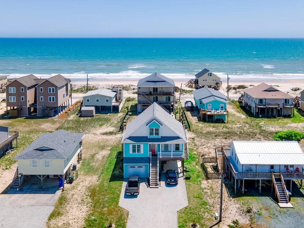 The hero image showcases a stunning blue beach house perched right on the sandy shores of Surf City, North Carolina. The house offers panoramic ocean views, with gentle waves and a wide, inviting stretch of sand just steps from the porch. Neighboring beach homes hint at a friendly seaside community, while the serene atmosphere and pastel sky suggest a perfect spot for relaxation and family fun.