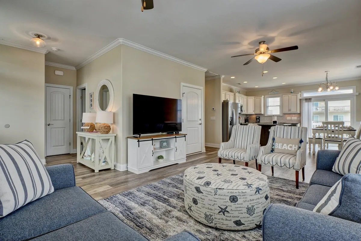 This image showcases a welcoming living area with a striped-cushion sofa, round ottoman with nautical design, TV on a media console, and a dining area with chandelier. Large windows allow bright coastal sunlight to fill the space, enhancing the cheerful beach-themed ambiance.
