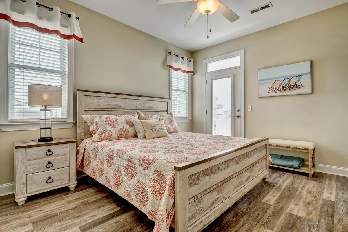 A serene bedroom featuring a bed with a plush decorative pillow, a nightstand with a lamp, a large window letting in natural light, and calming artwork on the walls. The space feels cozy and tranquil, ideal for rest and relaxation.