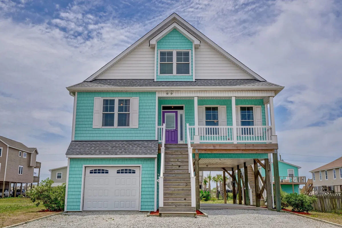 A bright exterior view of the house with turquoise siding and a cheerful purple door, a staircase ascending to a welcoming porch with white railings. The porch offers seating for enjoying sunset views and plenty of space to gather or watch the world go by.