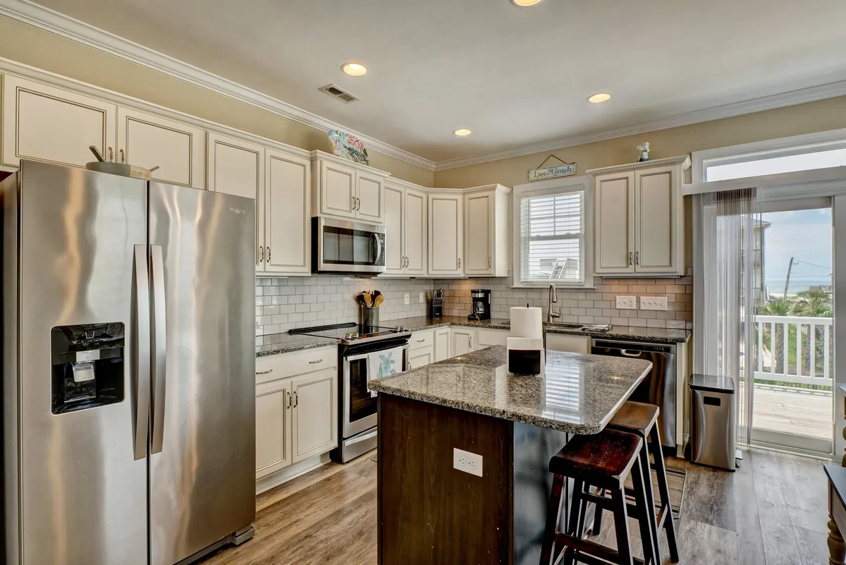 A modern kitchen with a stainless steel refrigerator, granite countertop, kitchen island, and a window offering a scenic view. The space is both functional and inviting, ideal for cooking, entertaining, or enjoying a morning coffee.