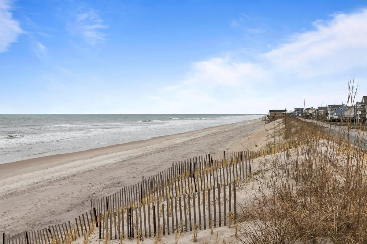 Expansive sandy beach with gentle dunes, ocean waves, and houses in the distance. The serene setting is ideal for long, relaxing walks with a dog along the shore.
