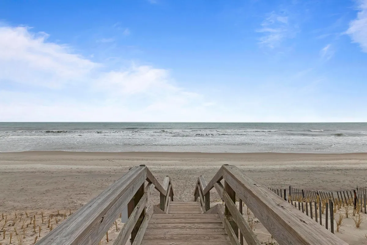Wooden steps lead down through dune grass and fencing to an expansive view of the ocean, its gentle waves meeting the sandy shore under a serene sky. The inviting pathway suggests immediate and convenient beach access just steps from home.