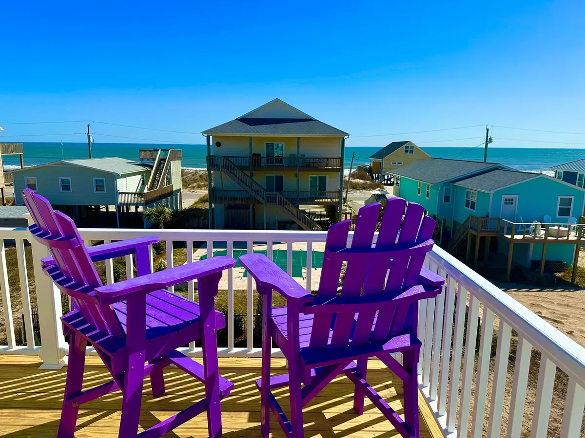 A tranquil view from a balcony with purple Adirondack chairs facing the ocean and colorful beach houses. The scene suggests a relaxing spot to enjoy coffee, ocean breezes, and memorable sunrises or sunsets.