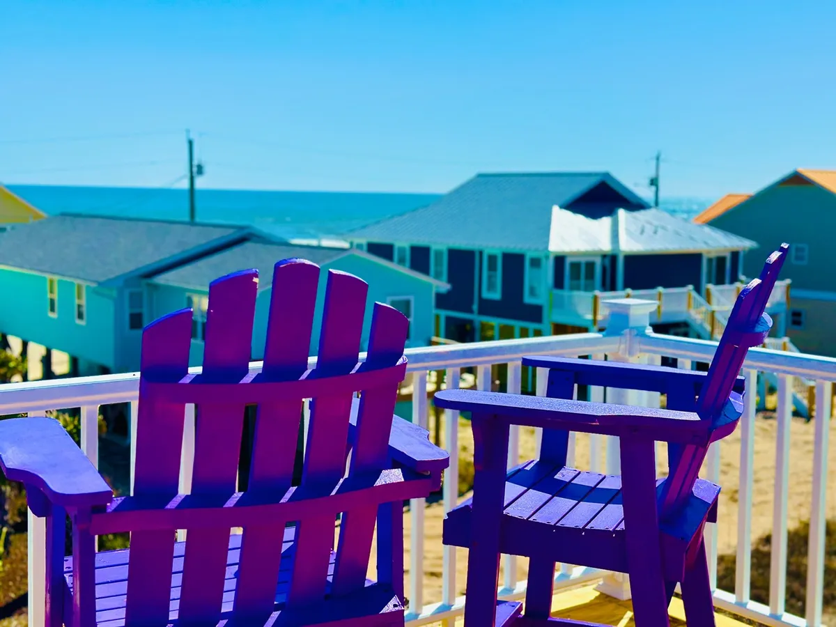A set of purple Adirondack chairs sits on a deck overlooking the ocean and neighboring colorful beach houses. The scene exudes an inviting, social atmosphere, ideal for group relaxation, sunset watching, and sharing drinks or stories together while enjoying the ocean breeze.