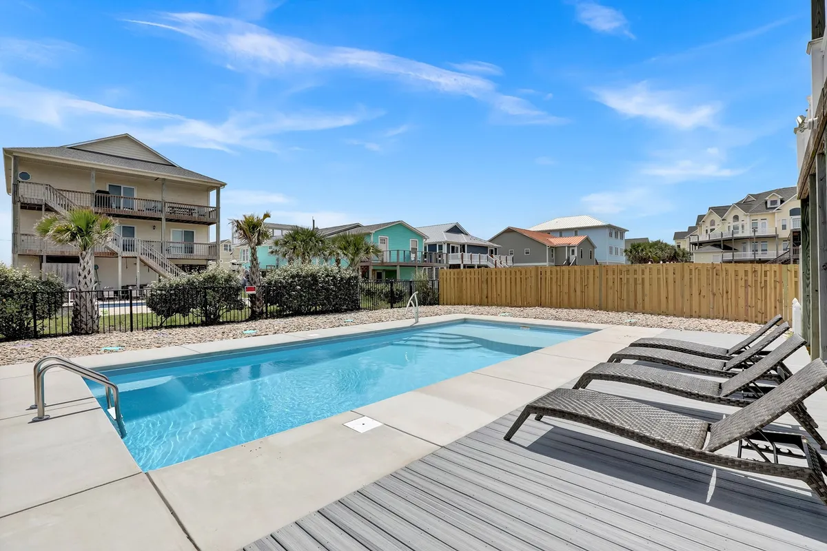 A fenced backyard featuring a swimming pool, lounge chairs, palm trees, and ample grassy space. The secure, open yard suggests a perfect spot for pets to play safely before heading to the beach.