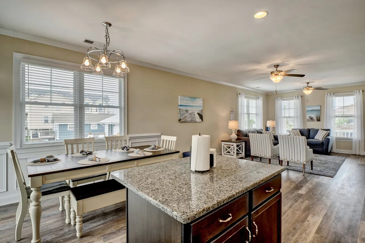 A spacious open-plan area featuring a dining table and kitchen island, with the living area and large windows in the background. Natural light streams in, emphasizing the seamless flow between kitchen, dining, and living spaces.