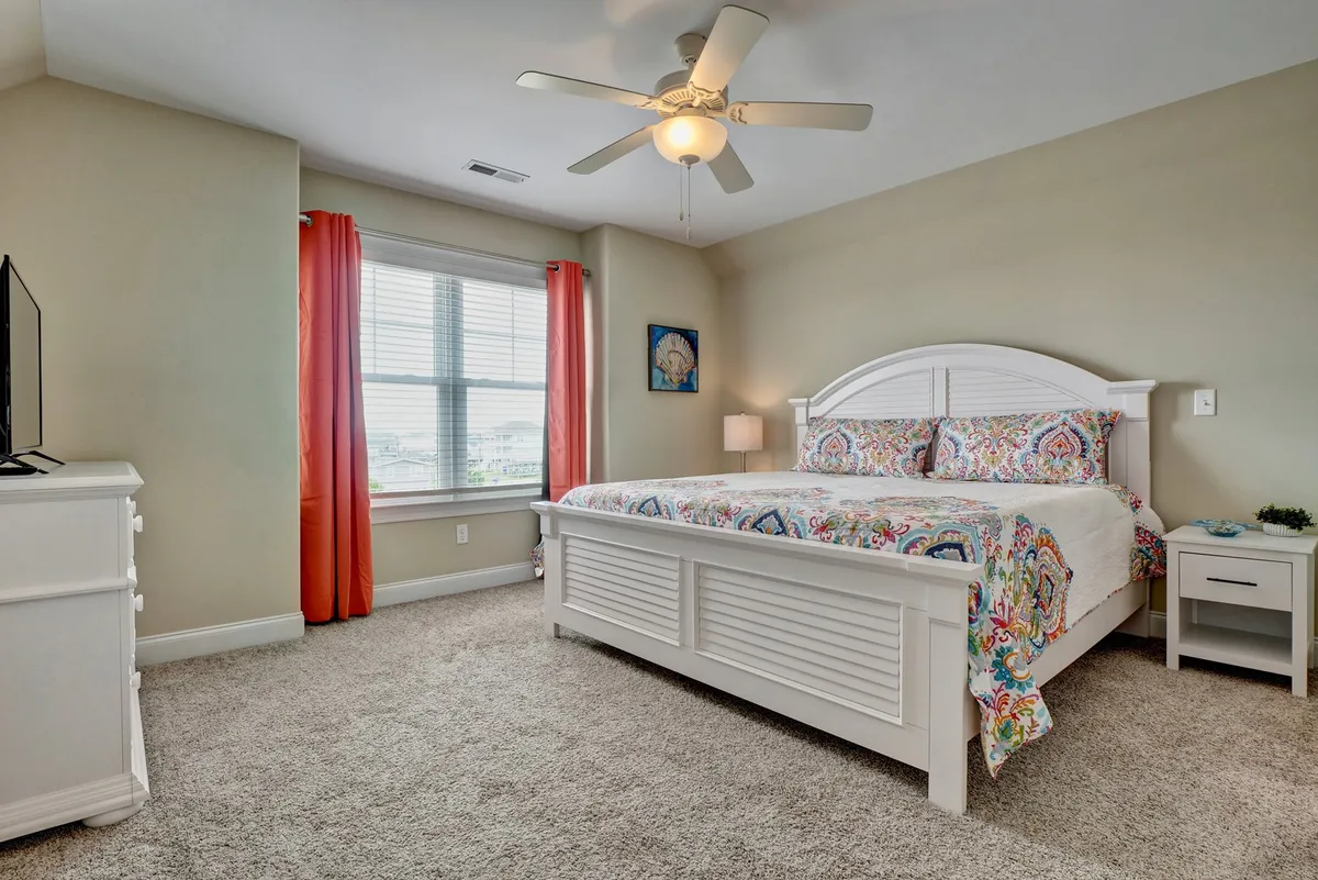 A cheerful bedroom with colorful bedding on a spacious bed, a large window that floods the room with natural light, a ceiling fan overhead, and a television positioned for convenient viewing. The decor feels inviting and perfect for unwinding after a beach day.