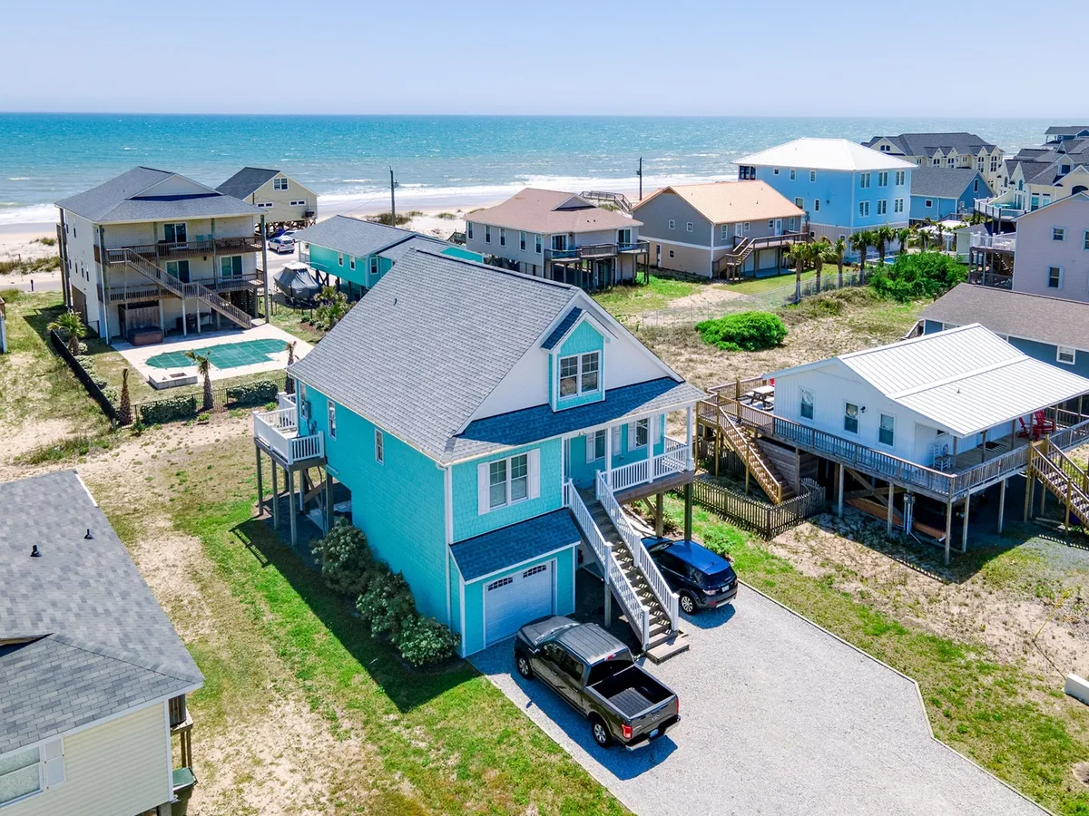 This image features a beachfront house with clear blue ocean in the background, surrounding houses, and vehicles parked in the driveway. The driveway and ample parking area are clearly visible, and the ocean is just steps away, setting a scene of easy access to beach adventures and sustainable travel.
