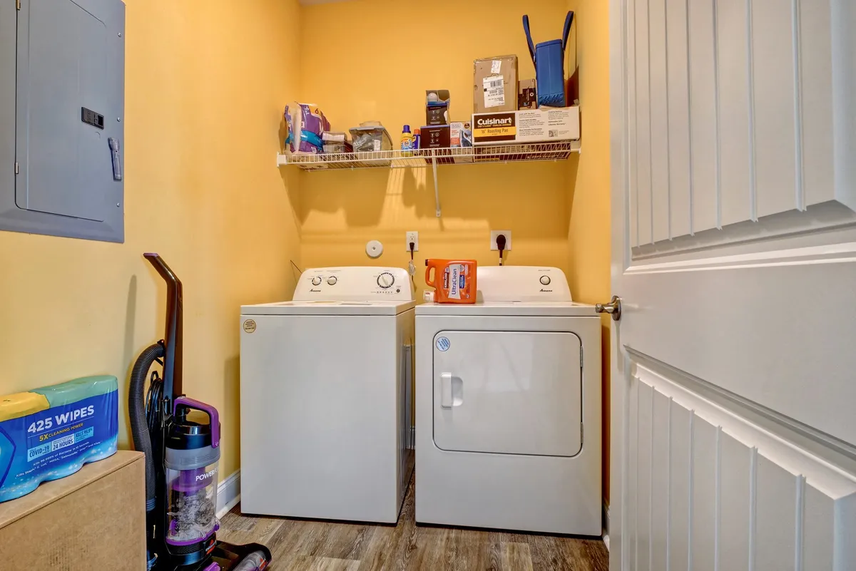 A bright and functional laundry area featuring a modern washing machine and dryer, shelves stocked with cleaning supplies, and a vacuum cleaner placed neatly nearby. The clean design emphasizes utility and convenience, making laundry days efficient and comfortable.