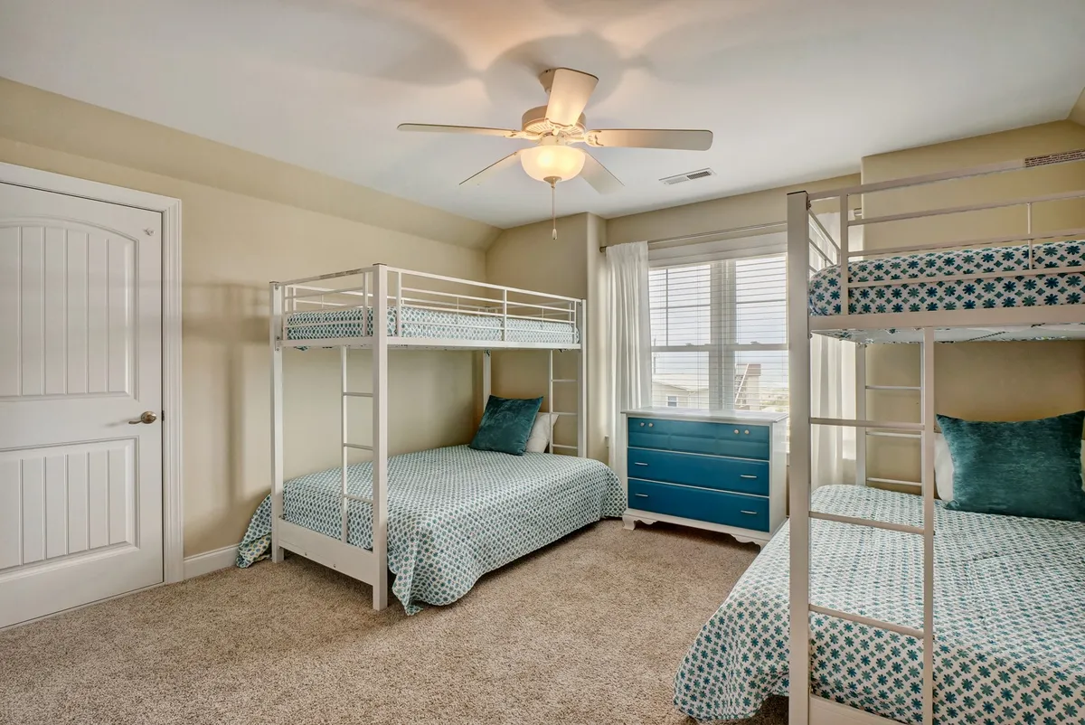 A playful bunk room with two sets of bunk beds, vibrant bedding, a blue dresser, and a large window. The room is filled with light and energy, ideal for kids or friends to enjoy sleepovers and fun nights together.